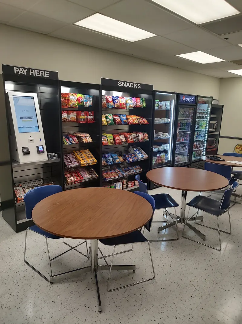 A micro market with a pay-here kiosk, snack shelves, drink coolers, and café tables